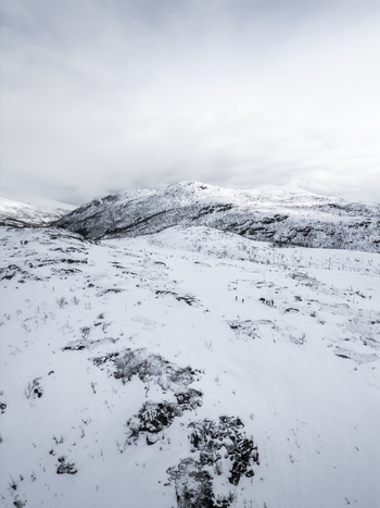 Aerial view of snow covered mountain peaks and rocky slopes under an overcast sky with people trekking near Ersfjord Troms Norway. Aerial view of snow covered mountain peaks and rocky slopes under an overcast sky with people trekking near Ersfjord Troms Norway.