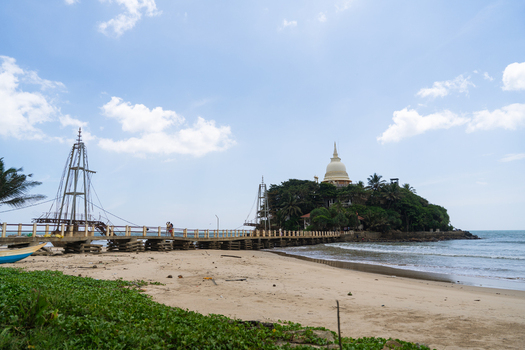 View of the Parevi Duwa Temple on a small island connected by a footbridge to the sandy beach under a blue sky Matara Southern Province Sri Lanka.