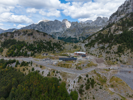 Aerial view of the Albanian Alps featuring a mountain lodge a winding road and dense pine forests under a cloudy sky in Shkoder Shkoder County Albania. Aerial view of the Albanian Alps featuring a mountain lodge a winding road and dense pine forests under a cloudy sky in Shkoder Shkoder County Albania.