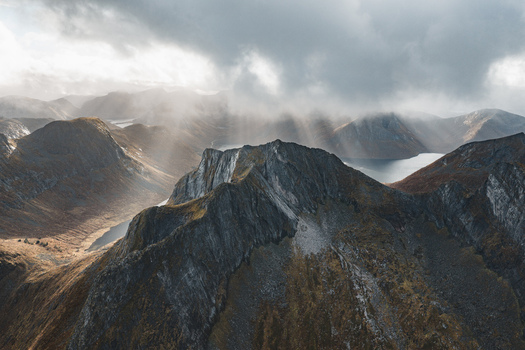 Aerial view of the dramatic mountain peaks and a lake nestled in the valley under a cloudy sky with sunbeams breaking through Senja Troms Norway.