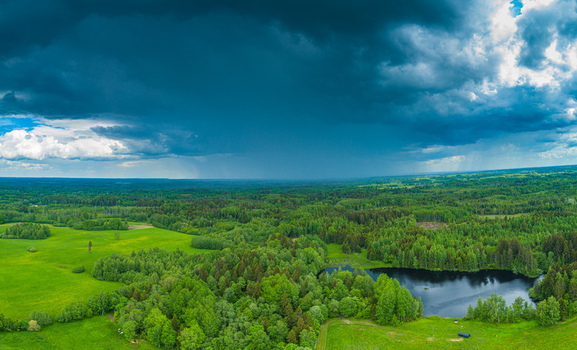 Aerial view of a lush green landscape with dense forests open fields and a small lake under a dramatic stormy sky in Latvia. Aerial view of a lush green landscape with dense forests open fields and a small lake under a dramatic stormy sky in Latvia.
