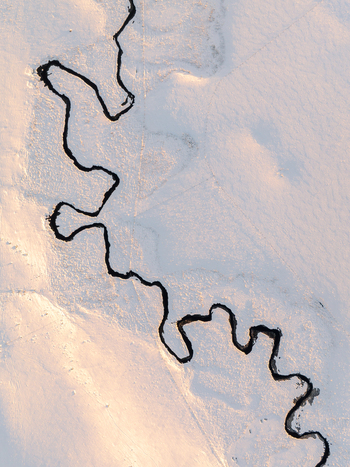 Aerial view of a dark meandering river carves a stark contrast through the pristine snow covered landscape under the soft glow of sunlight Nasbinals Occitanie France.