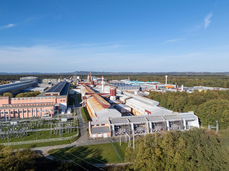 Aerial view of a large industrial complex with warehouses chimneys and an electrical substation surrounded by dense green forest under a clear blue sky in Slovenia.