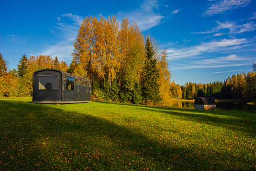 View of a modern dark colored cabin situated in a grassy field with autumn trees and a small barrel shaped structure near a pond in Latvia. View of a modern dark colored cabin situated in a grassy field with autumn trees and a small barrel shaped structure near a pond in Latvia.