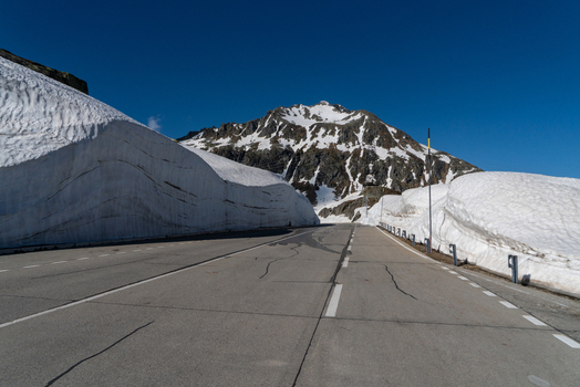 View of the Sankt Gotthard Pass road flanked by high snow walls leading towards a rocky mountain peak under a clear blue sky in Airolo Ticino Switzerland.
