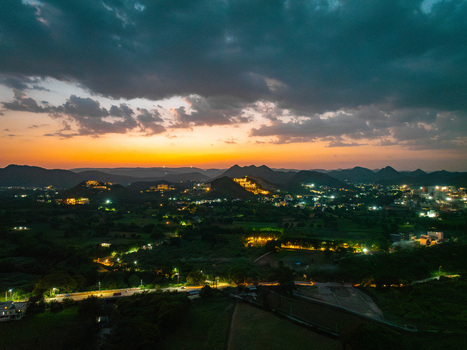Aerial view of the city at dusk with illuminated buildings nestled among rolling hills under a vibrant sunset sky in Udaipur Rajasthan India.
