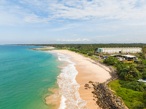 Aerial view of the sandy coastline with turquoise ocean waves industrial buildings and lush tropical vegetation in Peraliya Southern Province Sri Lanka. Aerial view of the sandy coastline with turquoise ocean waves industrial buildings and lush tropical vegetation in Peraliya Southern Province Sri Lanka.
