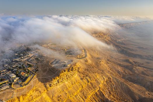 Aerial view of the Makhtesh Ramon crater with low hanging clouds spilling over the rugged desert cliffs at golden hour in Mitzpe Ramon South District Israel. Aerial view of the Makhtesh Ramon crater with low hanging clouds spilling over the rugged desert cliffs at golden hour in Mitzpe Ramon South District Israel.