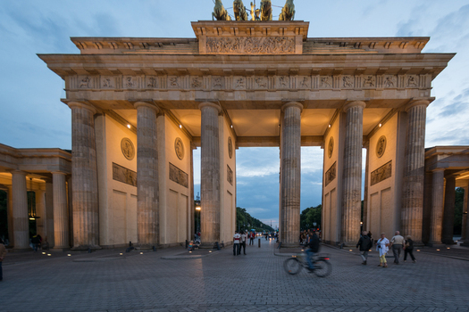View of the Brandenburg Gate with its neoclassical columns illuminated at dusk featuring people walking and a cyclist on the cobblestone square in Berlin Berlin Germany.