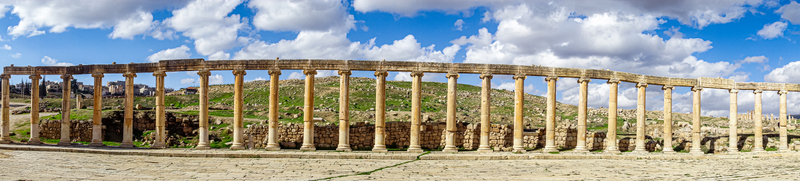 View of the ancient Roman colonnade of the Oval Plaza in Jerash featuring weathered stone pillars against rolling green hills and a bright blue sky in Jerash Jerash Governorate Jordan. View of the ancient Roman colonnade of the Oval Plaza in Jerash featuring weathered stone pillars against rolling green hills and a bright blue sky in Jerash Jerash Governorate Jordan.