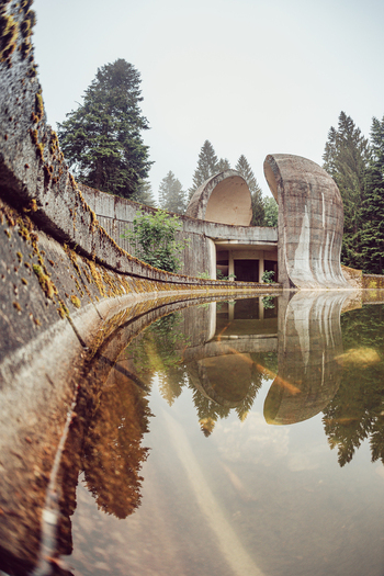 View of the brutalist Korcanica Memorial Zone reflecting in a still pool surrounded by pine trees in Sanski Most Federation of Bosnia Bosnia and Herzegovina. View of the brutalist Korcanica Memorial Zone reflecting in a still pool surrounded by pine trees in Sanski Most Federation of Bosnia Bosnia and Herzegovina.