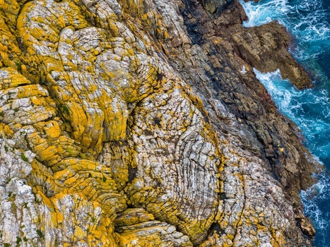 Aerial view of the rugged cliffs at Silence Beach Viewpoint with yellow lichen and crashing blue waves in Cudillero Asturias Spain.