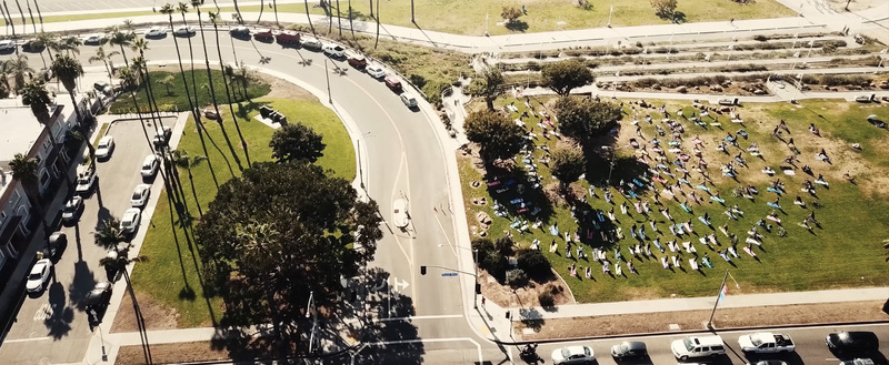 Aerial view of a large group of people practicing yoga on colorful mats in a sunny public park surrounded by palm trees and city streets in Los Angeles California United States.