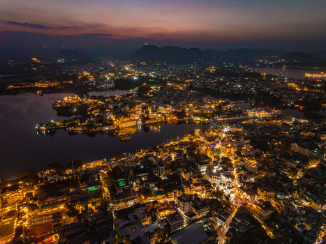 Aerial view of the illuminated cityscape at night with glowing lights of the City Palace and buildings reflecting on the calm waters of Lake Pichola Udaipur Rajasthan India.