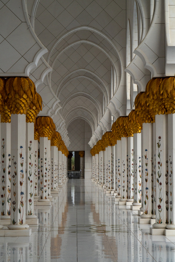 View of the ornate colonnade featuring white marble columns with floral inlays and golden palm capitals under pointed arches in Abu Dhabi Abu Dhabi United Arab Emirates. View of the ornate colonnade featuring white marble columns with floral inlays and golden palm capitals under pointed arches in Abu Dhabi Abu Dhabi United Arab Emirates.