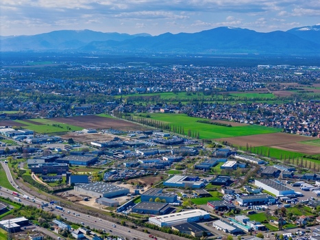 Aerial view of the sprawling industrial park contrasting with the vibrant green fields against a backdrop of majestic mountains Sausheim Grand Est France.