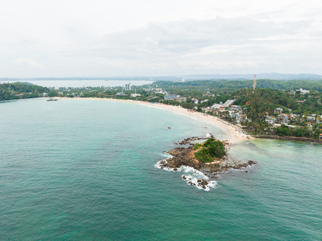 Aerial view of the crescent shaped Mirissa Beach and Parrot Rock surrounded by turquoise waters and lush tropical vegetation in Mirissa Southern Province Sri Lanka. Aerial view of the crescent shaped Mirissa Beach and Parrot Rock surrounded by turquoise waters and lush tropical vegetation in Mirissa Southern Province Sri Lanka.