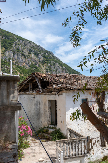 View of an old stone house with a damaged tiled roof and white walls set against a steep rocky mountain under a blue sky in Qeparo Fshat Vlore County Albania. View of an old stone house with a damaged tiled roof and white walls set against a steep rocky mountain under a blue sky in Qeparo Fshat Vlore County Albania.