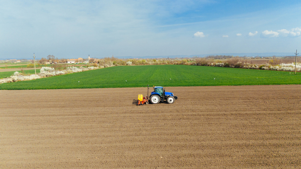 Aerial view of a blue tractor cutting through the vast expanse of brown and green fields under a partly cloudy sky Sremska Mitrovica Vojvodina Serbia. Aerial view of a blue tractor cutting through the vast expanse of brown and green fields under a partly cloudy sky Sremska Mitrovica Vojvodina Serbia.