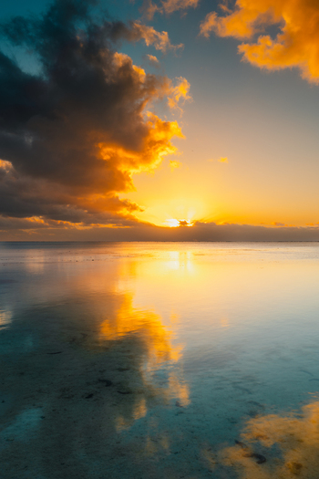 View of the sunset over the calm lagoon of Aitutaki with golden clouds reflecting on the water in Cook Islands. View of the sunset over the calm lagoon of Aitutaki with golden clouds reflecting on the water in Cook Islands.