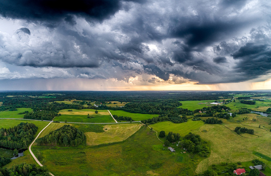 Aerial view of a vast rural landscape featuring lush green fields dense forests and winding roads under a dramatic stormy sky with heavy rain clouds in Latvia. Aerial view of a vast rural landscape featuring lush green fields dense forests and winding roads under a dramatic stormy sky with heavy rain clouds in Latvia.