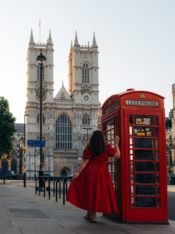 View of a woman in a vibrant red dress pauses by a classic red telephone box with the majestic Westminster Abbey rising in the background London England United Kingdom. View of a woman in a vibrant red dress pauses by a classic red telephone box with the majestic Westminster Abbey rising in the background London England United Kingdom.