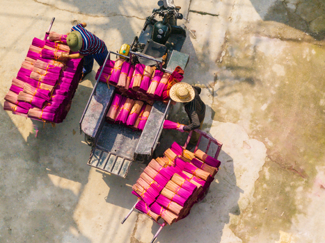 Aerial view of workers loading bundles of bright pink incense sticks onto a motorcycle cart on a concrete surface in Vietnam. Aerial view of workers loading bundles of bright pink incense sticks onto a motorcycle cart on a concrete surface in Vietnam.