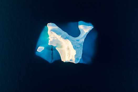 Aerial view of a bright white iceberg floating in the deep blue waters of the Arctic Ocean illuminated by soft sunlight Greenland. Aerial view of a bright white iceberg floating in the deep blue waters of the Arctic Ocean illuminated by soft sunlight Greenland.
