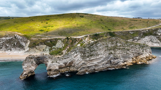 Aerial view of Durdle Door natural limestone arch on the Jurassic Coast with rocky cliffs green hills and sea under a cloudy sky in Wareham England United Kingdom. Aerial view of Durdle Door natural limestone arch on the Jurassic Coast with rocky cliffs green hills and sea under a cloudy sky in Wareham England United Kingdom.