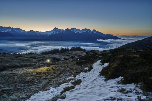 View of Valmalenco at dawn with a hiker using a headlamp on a snowy mountain trail above a sea of clouds in Lombardy Italy. View of Valmalenco at dawn with a hiker using a headlamp on a snowy mountain trail above a sea of clouds in Lombardy Italy.
