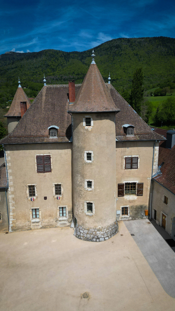 Aerial view of the historic Chateau De Vesancy featuring a prominent round tower and tiled roofs against lush green hills in Vesancy Auvergne Rhone Alpes France. Aerial view of the historic Chateau De Vesancy featuring a prominent round tower and tiled roofs against lush green hills in Vesancy Auvergne Rhone Alpes France.