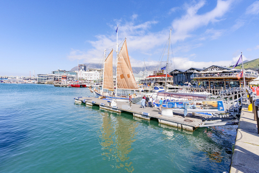 View of a traditional sailboat with brown sails docked at the Victoria and Alfred Waterfront with people walking on the pier in Cape Town Western Cape South Africa. View of a traditional sailboat with brown sails docked at the Victoria and Alfred Waterfront with people walking on the pier in Cape Town Western Cape South Africa.