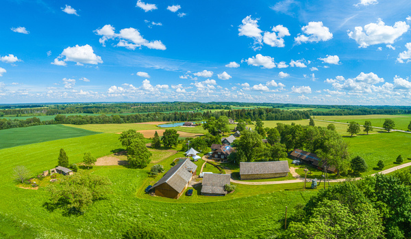 Aerial view of a rural farmstead with traditional wooden buildings surrounded by lush green fields trees and a small pond under a bright blue sky in Latvia. Aerial view of a rural farmstead with traditional wooden buildings surrounded by lush green fields trees and a small pond under a bright blue sky in Latvia.