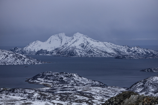 View of snow capped mountain peaks rising above a calm fjord under a moody winter sky Kvaloya Troms Norway.