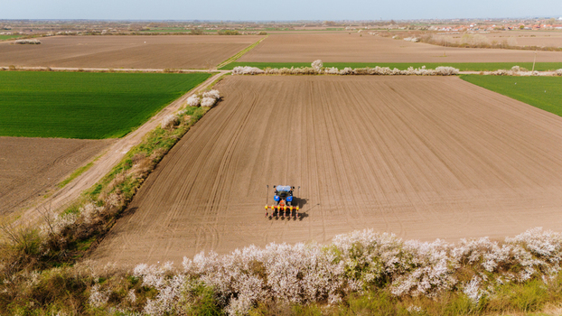 Aerial view of a tractor working the soil creating patterns of dark earth against the bright green fields bordered by blooming trees Sremska Mitrovica Vojvodina Serbia. Aerial view of a tractor working the soil creating patterns of dark earth against the bright green fields bordered by blooming trees Sremska Mitrovica Vojvodina Serbia.