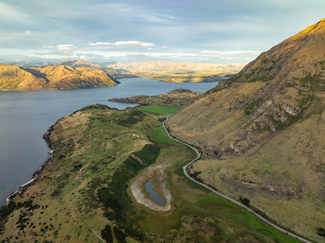 Aerial view of the winding road snaking through the rugged grassy hills alongside the tranquil lake waters reflecting the soft light of the sky Glendhu Bay Otago Region New Zealand. Aerial view of the winding road snaking through the rugged grassy hills alongside the tranquil lake waters reflecting the soft light of the sky Glendhu Bay Otago Region New Zealand.