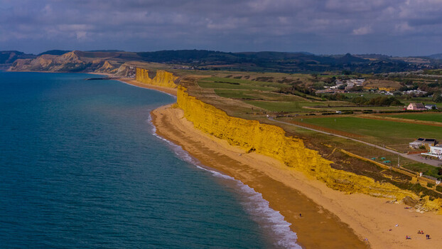 Aerial view of the golden sandstone cliffs of West Bay meeting the turquoise sea with a sandy beach and green coastal fields under a cloudy sky in West Bay England United Kingdom. Aerial view of the golden sandstone cliffs of West Bay meeting the turquoise sea with a sandy beach and green coastal fields under a cloudy sky in West Bay England United Kingdom.
