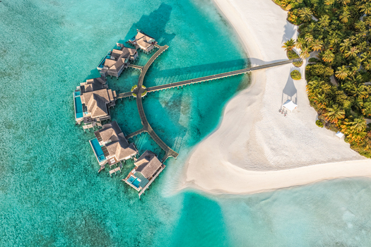 Aerial view of overwater bungalows with private pools connected by a wooden jetty to a white sand beach and lush tropical vegetation near Kudahuvadhoo Dhaalu Atoll Maldives. Aerial view of overwater bungalows with private pools connected by a wooden jetty to a white sand beach and lush tropical vegetation near Kudahuvadhoo Dhaalu Atoll Maldives.