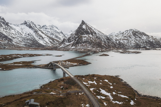 Aerial view of Flakstadbruene Bridge slicing through the icy waters contrasting with the snow capped peaks under a muted sky Flakstadbruene Nordland Norway. Aerial view of Flakstadbruene Bridge slicing through the icy waters contrasting with the snow capped peaks under a muted sky Flakstadbruene Nordland Norway.