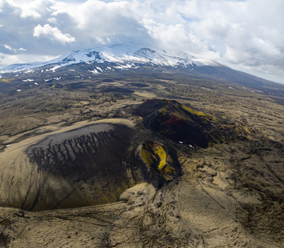 Aerial view of a dramatic volcanic landscape with a snow capped mountain peak in the background featuring rugged lava fields craters and mossy terrain in Iceland.
