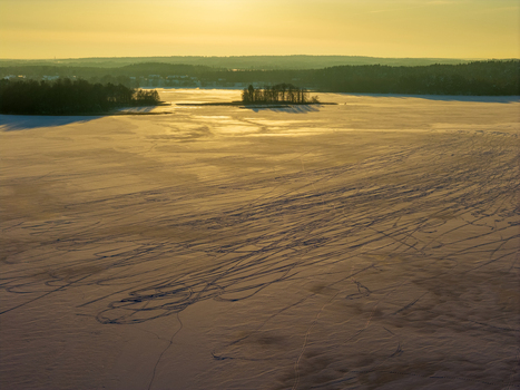 Aerial view of a frozen lake covered in snow with vehicle tracks under a golden sunset sky and distant forests in Lithuania. Aerial view of a frozen lake covered in snow with vehicle tracks under a golden sunset sky and distant forests in Lithuania.