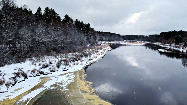 Aerial view of a river flowing through a snow covered pine forest under an overcast sky in Latvia. Aerial view of a river flowing through a snow covered pine forest under an overcast sky in Latvia.