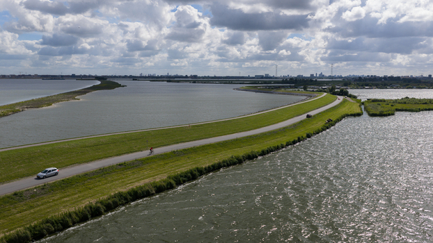 Aerial view of a dike road separating water bodies with a car and a cyclist on the road lush green grass and a distant city skyline in Uitdam Netherlands.