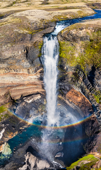 Aerial view of Haifoss waterfall cascading over basalt columns with a vibrant rainbow in the mist surrounded by mossy green terrain and rocky cliffs in Iceland. Aerial view of Haifoss waterfall cascading over basalt columns with a vibrant rainbow in the mist surrounded by mossy green terrain and rocky cliffs in Iceland.