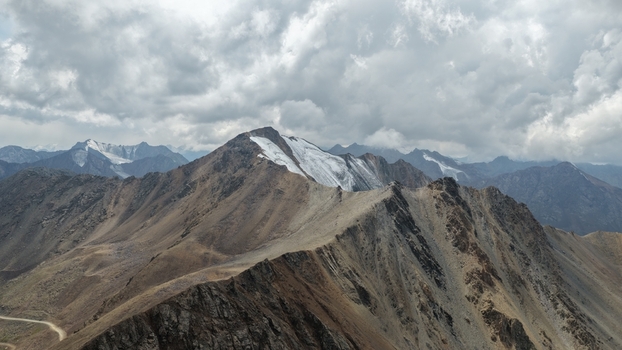 Aerial view of the rugged Enilchek mountain range with snow capped peaks rocky slopes and dramatic cloudy skies in Enilchek Issyk Kul Region Kyrgyzstan. Aerial view of the rugged Enilchek mountain range with snow capped peaks rocky slopes and dramatic cloudy skies in Enilchek Issyk Kul Region Kyrgyzstan.
