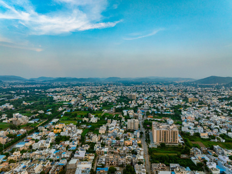 Aerial view of the sprawling cityscape of Udaipur featuring dense residential buildings lush green patches and distant hills under a vast blue sky in Udaipur Rajasthan India. Aerial view of the sprawling cityscape of Udaipur featuring dense residential buildings lush green patches and distant hills under a vast blue sky in Udaipur Rajasthan India.