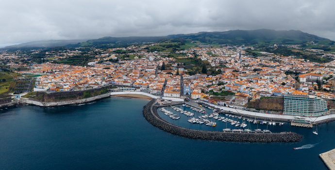 Aerial view of the historic coastal city featuring orange roofed houses a harbor filled with boats and lush green hills under a cloudy sky in Angra do Heroismo Azores Portugal.