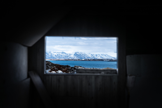 View of the interior of an abandoned farmhouse looking out through a window at snow capped mountains and the blue sea in Hafnarfjordur Hafnarfjardarkaupstad Iceland. View of the interior of an abandoned farmhouse looking out through a window at snow capped mountains and the blue sea in Hafnarfjordur Hafnarfjardarkaupstad Iceland.