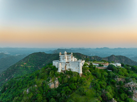Aerial view of Sajjangarh Palace perched on a hilltop surrounded by lush green forests and rolling hills under a soft golden hour sky in Udaipur Rajasthan India.