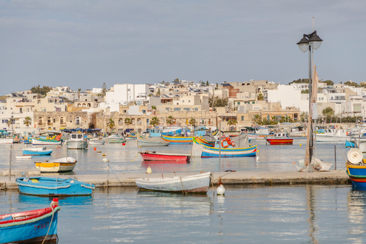 View of vibrant fishing boats bobbing gently on the cerulean water mirroring the bright buildings under the soft sky Marsaxlokk Malta. View of vibrant fishing boats bobbing gently on the cerulean water mirroring the bright buildings under the soft sky Marsaxlokk Malta.